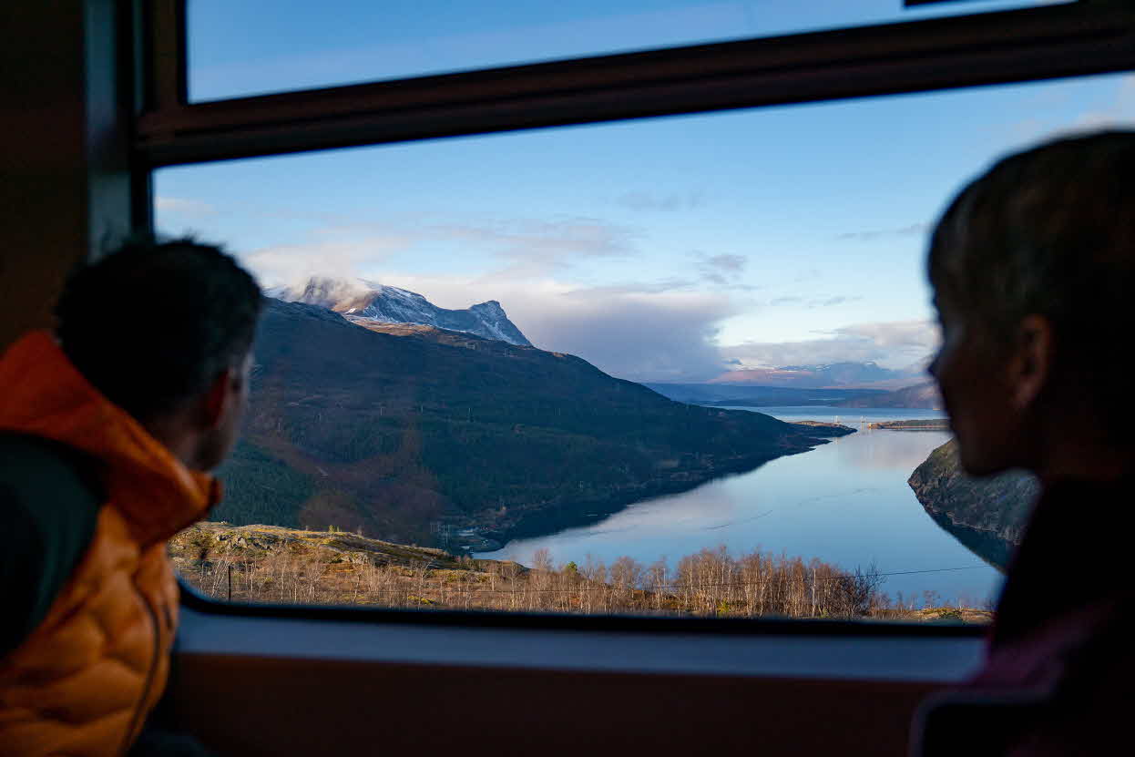 To personer sitter ovenfor hverandre ombord på Arctic Train og kikker ut mot Hålogalandsbrua og Rombaksfjorden