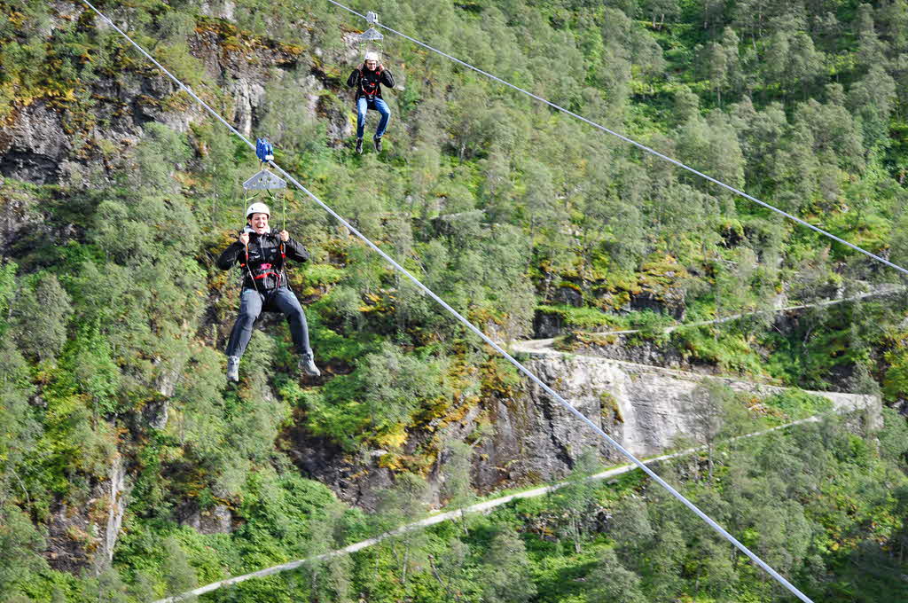 Two people flying down the Flåm Zipline with the hairpin turns of Rallarvegen behind.