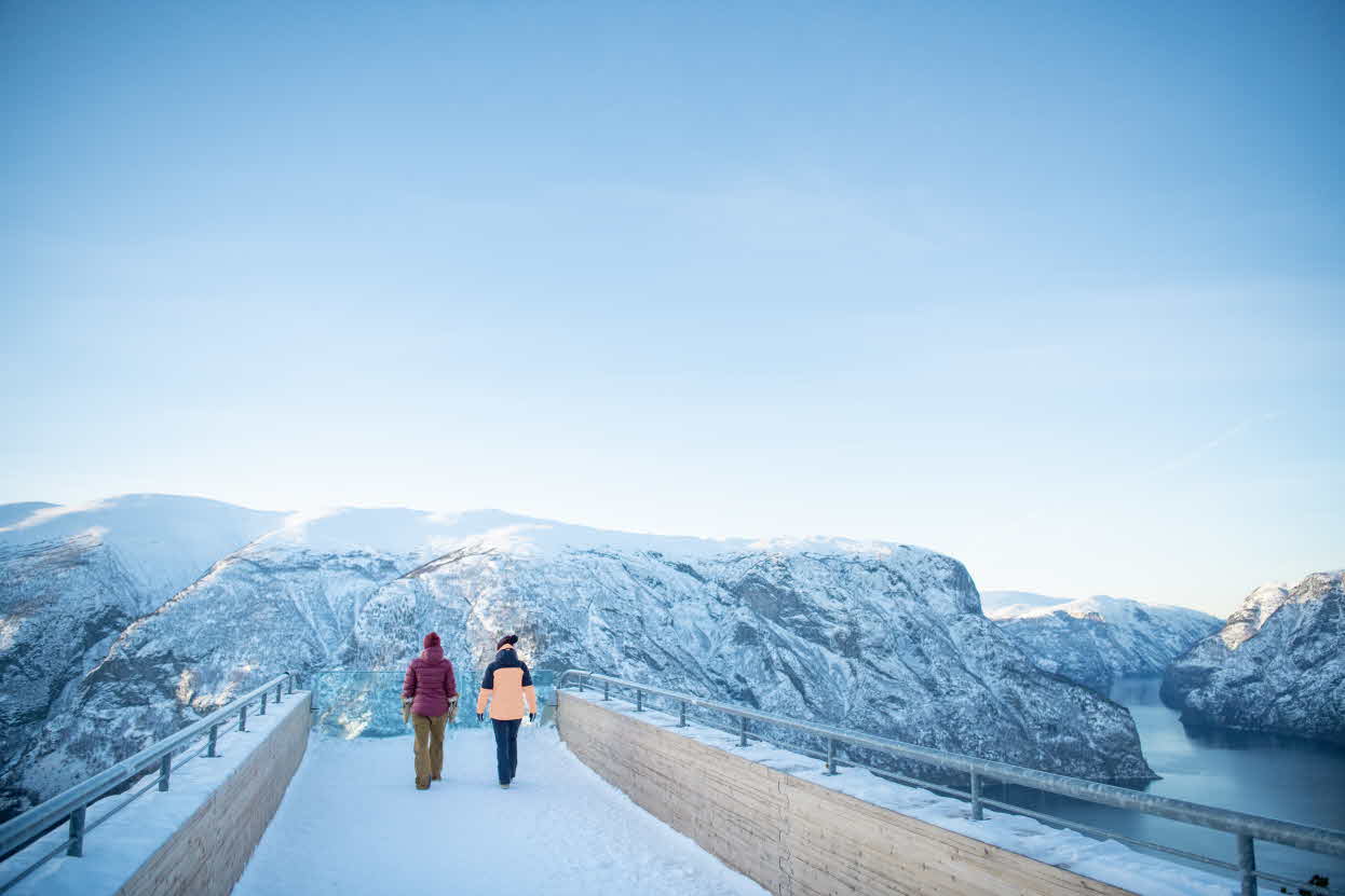 To personer i fargerik vinterbekledning går utover utsiktspunktet Stegastein ein fin og snørik vinterdag
