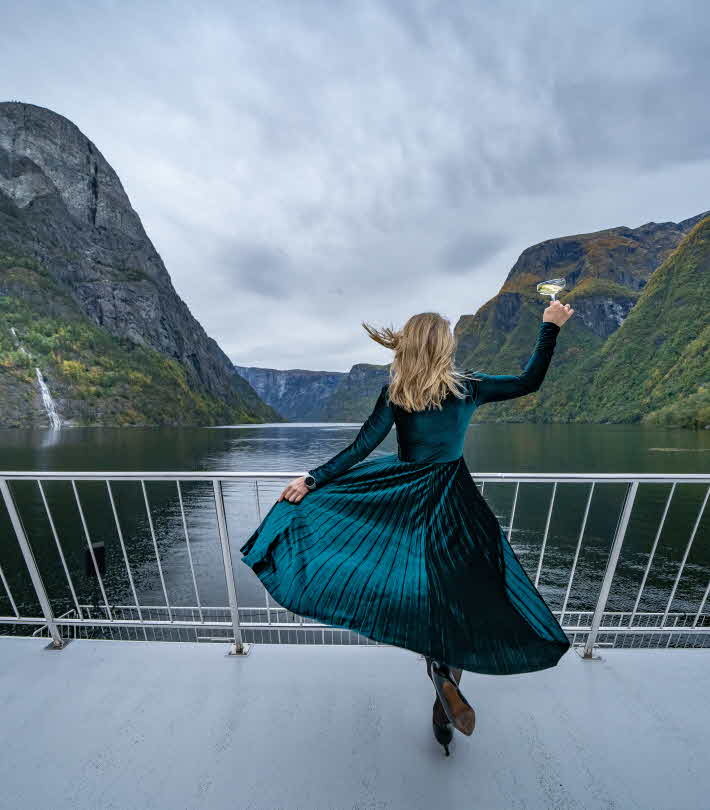 Kvinne med blafrande grønn kjole står med ryggen til i baugen og hevar eit glass mot Nærøyfjorden