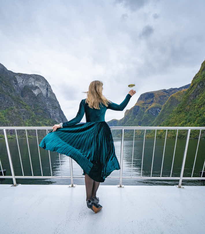 Ei kvinne poserer i grøn kjole med sjampagneglas i handa på ein båt på Nærøyfjorden