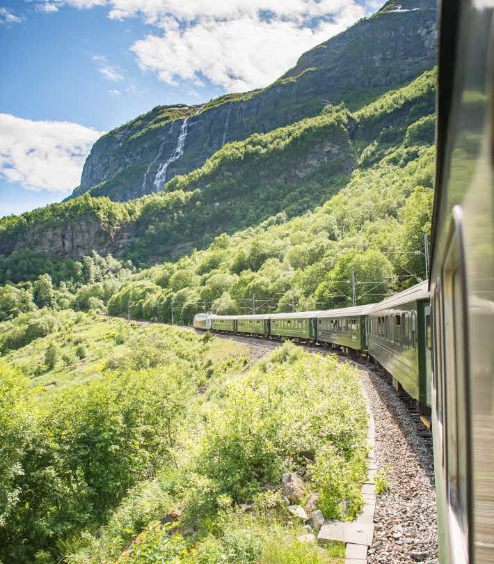 Passasjer om bord Flåmsbana tek bilete ut av vindauge av den frodige Flåmsdalen der ein ser heile toget