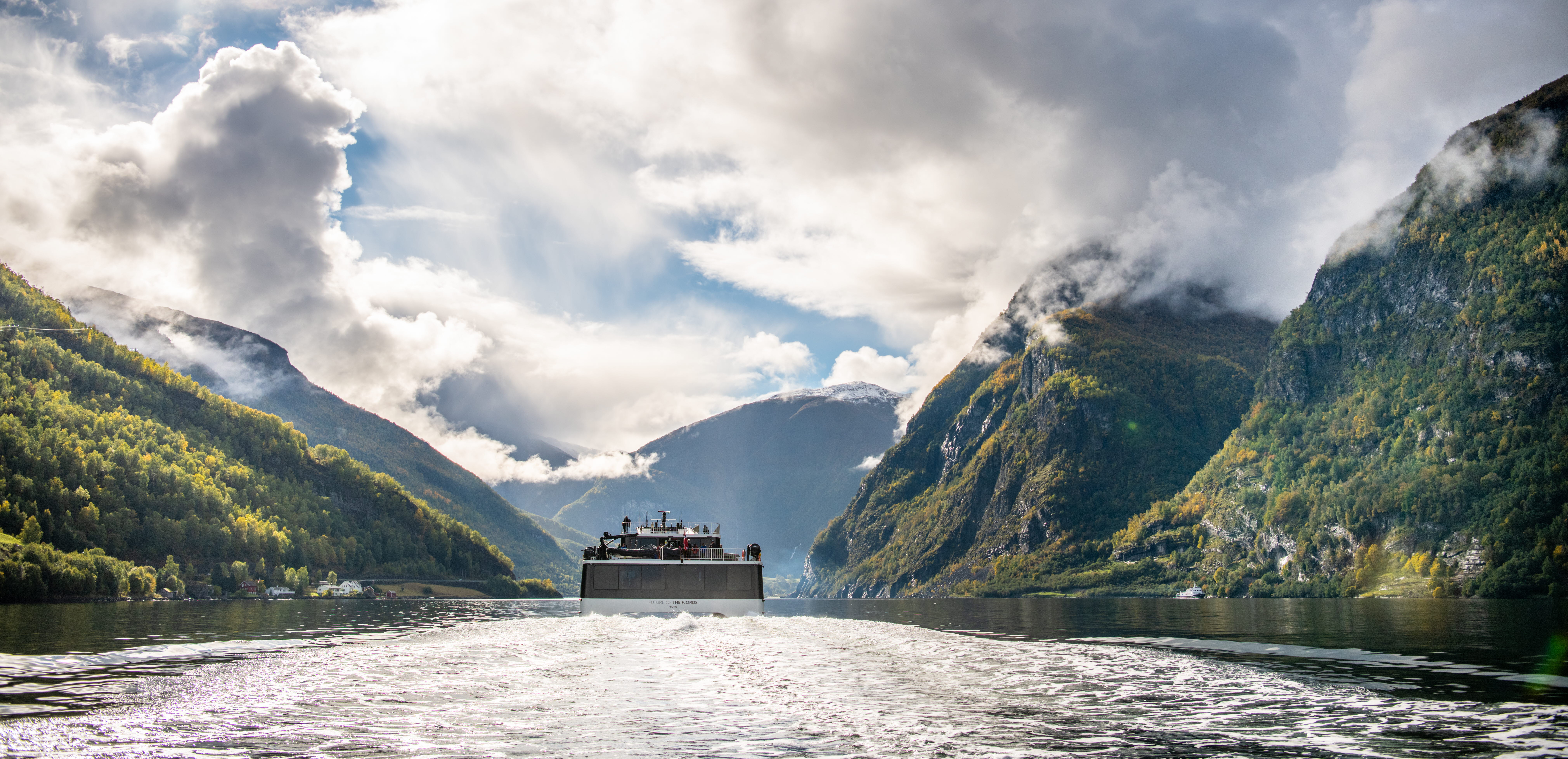 An electric catamaran seen sailing on the Nærøyfjord seen from behind. Mountains partly covered in clouds.
