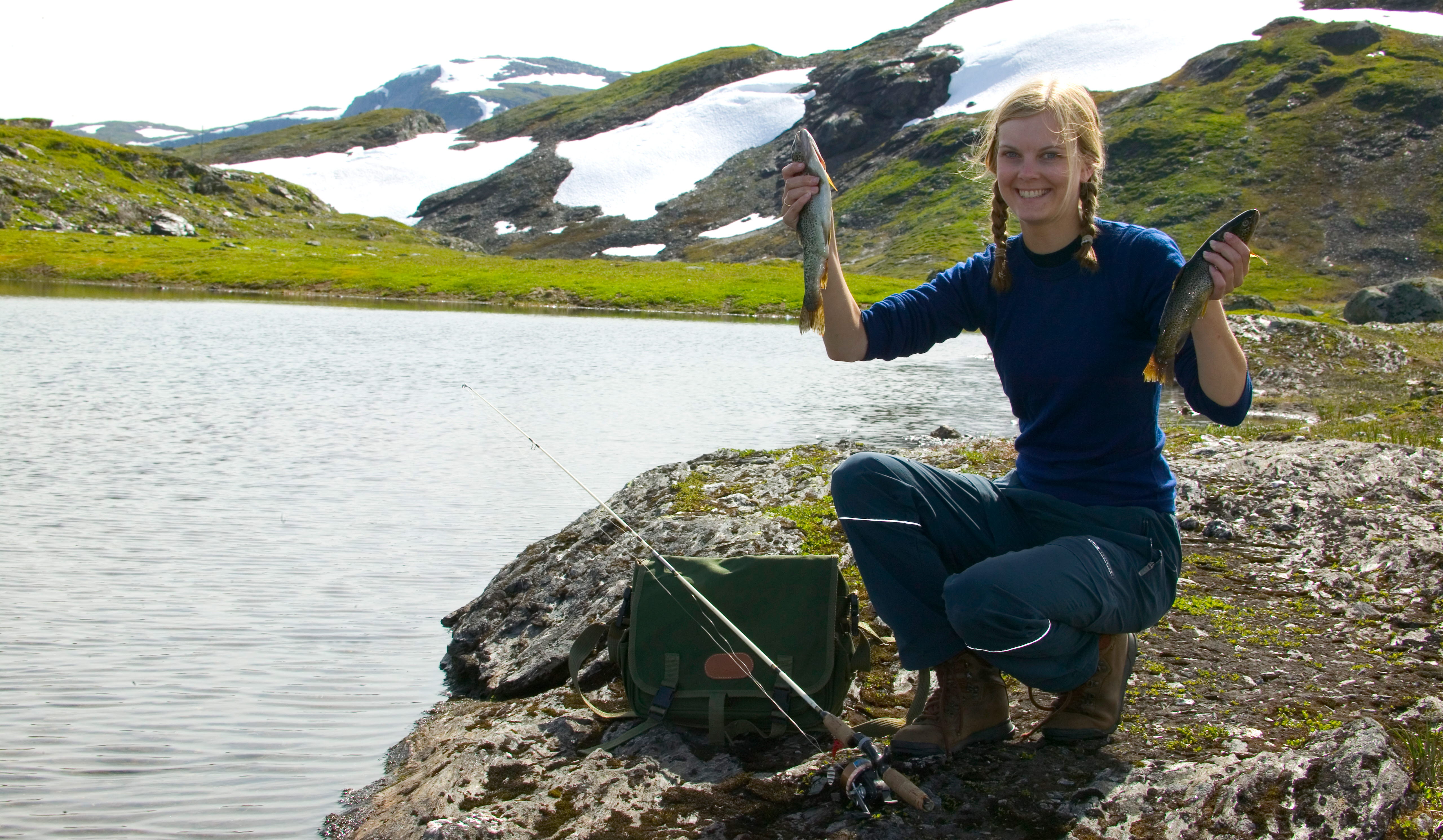 A teenage girl holding up two fish by a mountain lake with patches of snow in the background.